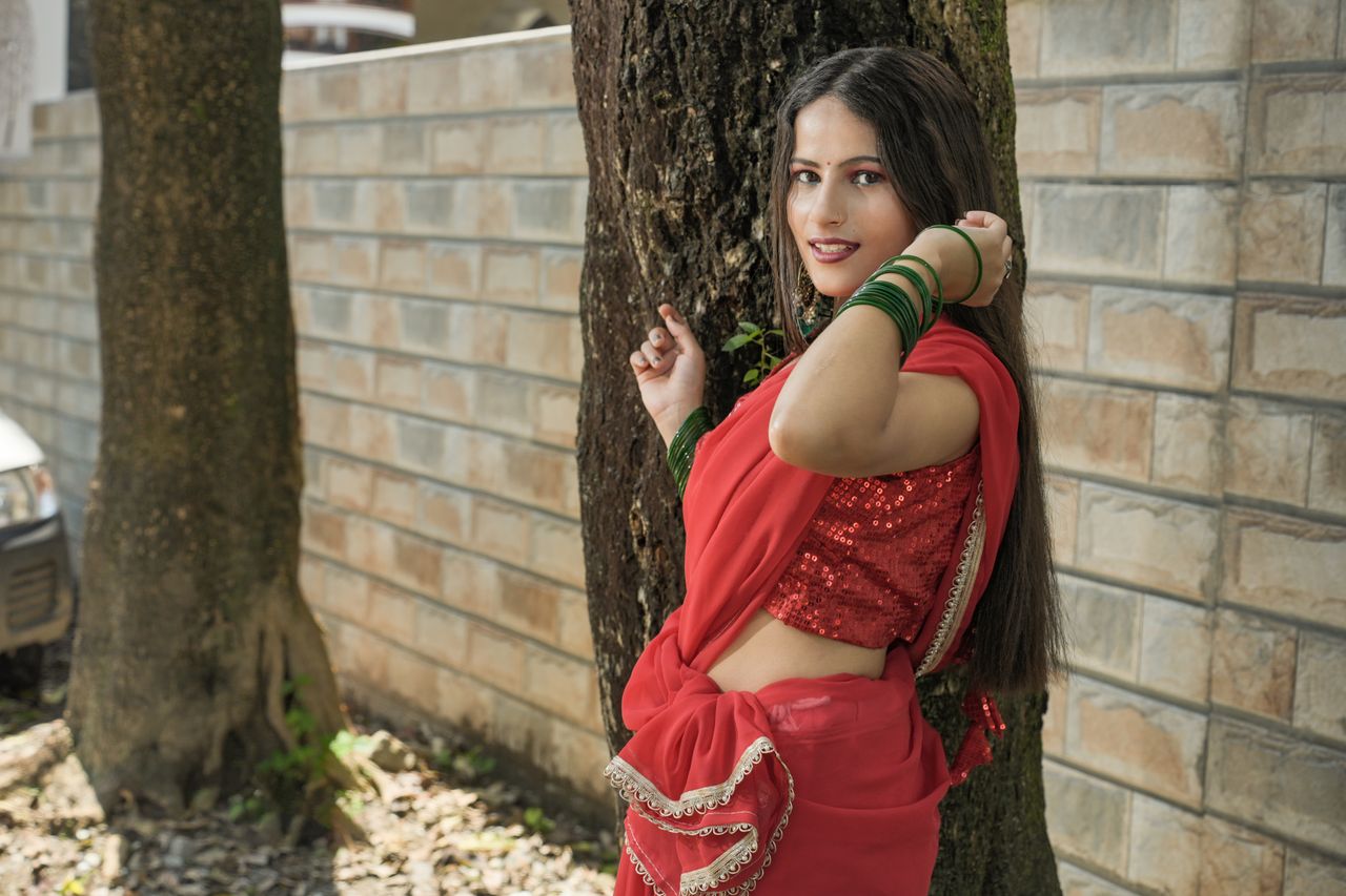 Elegant woman wearing a red sari poses confidently against a tree outdoors.